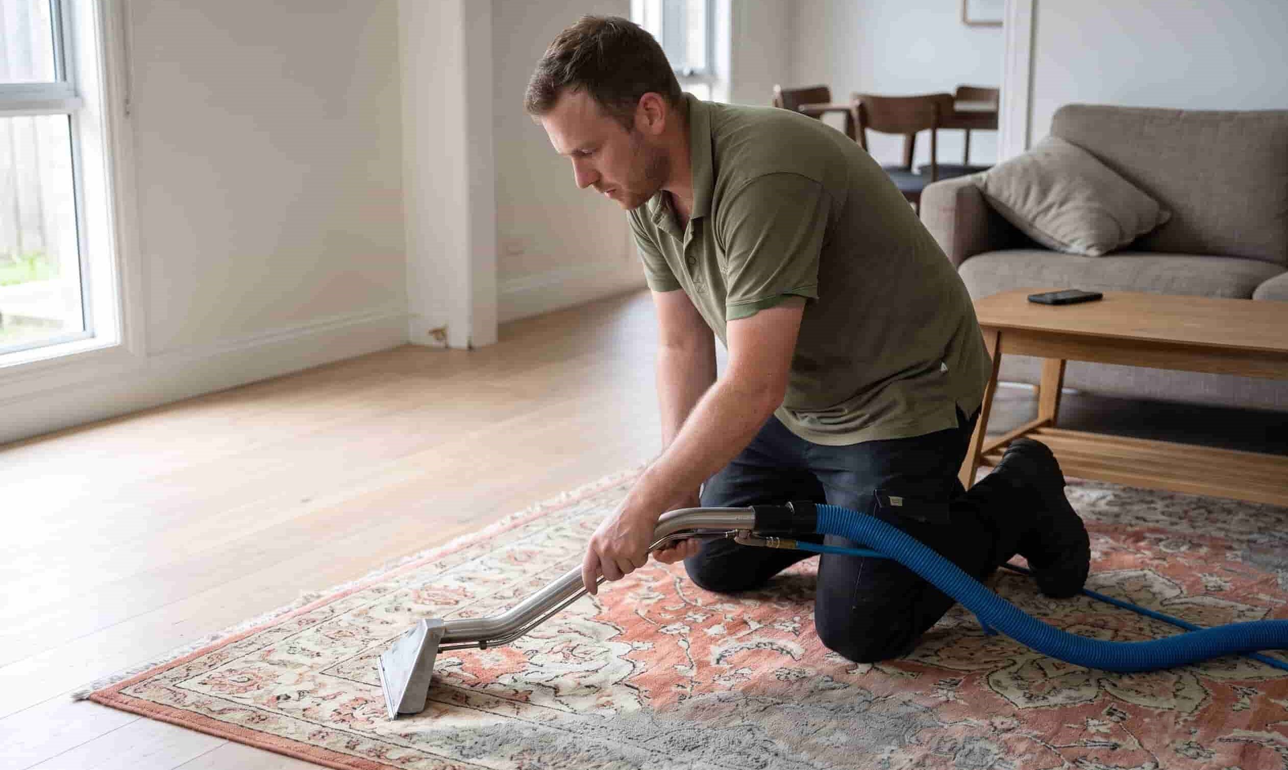 A professional cleaner kneeling and using a specialized extraction wand to clean a patterned area rug in a modern, well-lit living room.